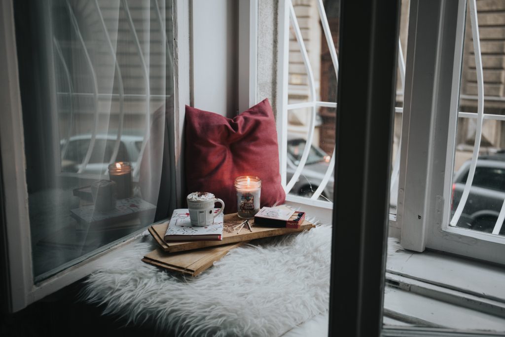 Auf dem Fensterbrett ist eine gemütliche Ecke für dich hergerichtet: Mit Kissen, heißer Schokolade, Kerze und Bücher. Feier dich und mach es deinen neuen Entscheidungen gemütlich bei dir!
